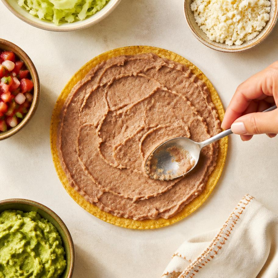 A hand spreading refried beans across a flat taco shell, surrounded by bowls of toppings